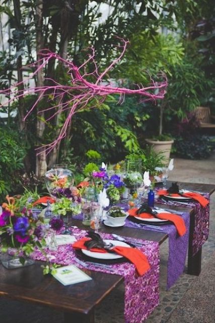 a bold fall wedding tablescape with fuchsia, pink and purple runners, orange napkins, purple, fuchsia and orange blooms and greenery and pink branches over the table