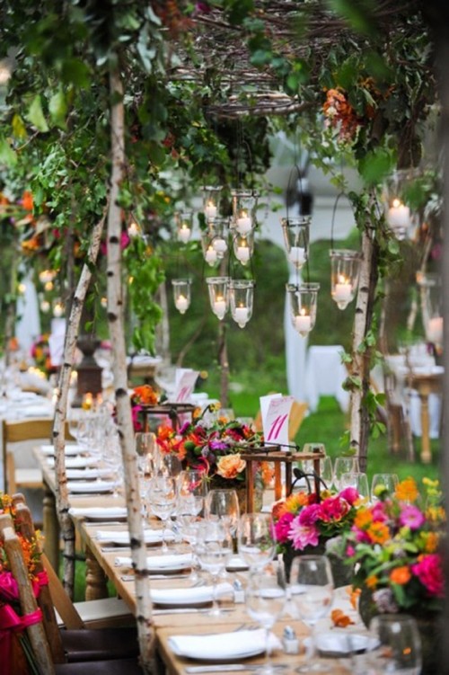 a rustic wedding tablescape with super bold blooms and greenery, candle lanterns, white porcelain and napkins, hanging candleholders and greenery over the table
