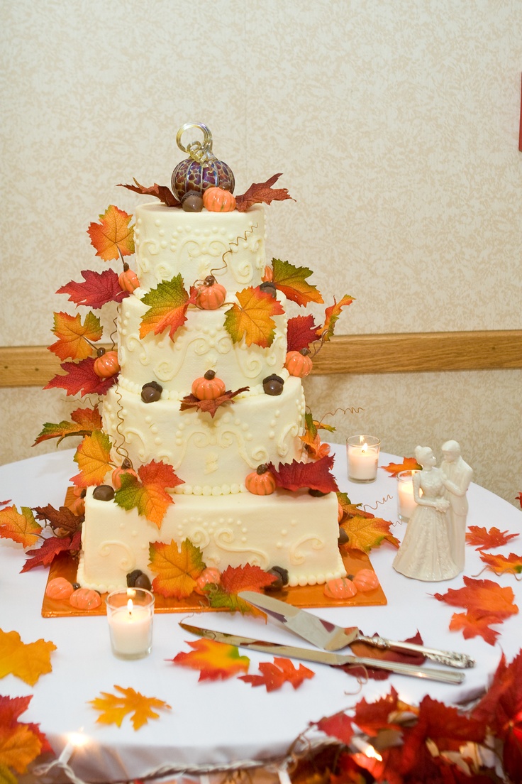 A patterned square and round wedding cake with fall leaves and pumpkins and a fun pumpkin topper