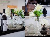 a wedding tablescape with neutral bottles with greenery and blooms and a wine bottle with a black table number