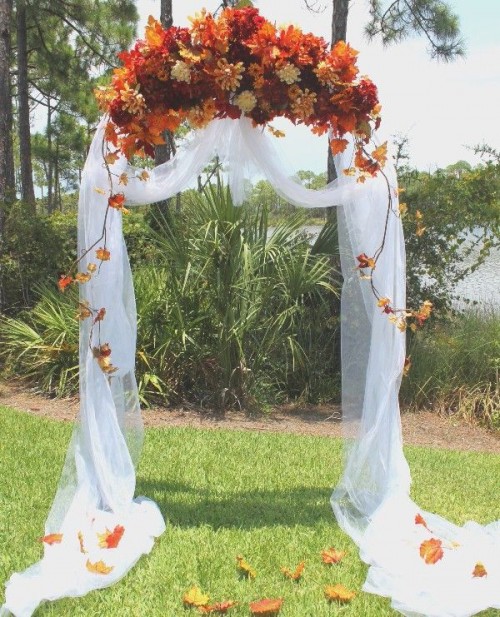 an outdoor fall wedding arch decorated with white fabric and lush and bright fall blooms and leaves