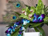 a bright wedding centerpiece with purple and green orchids and peacock feathers on a tall stand