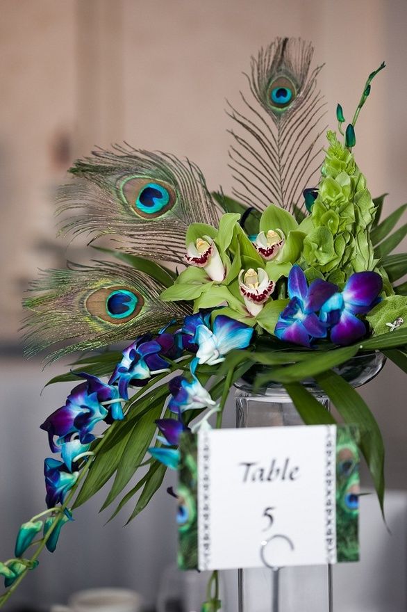 A bright wedding centerpiece with purple and green orchids and peacock feathers on a tall stand