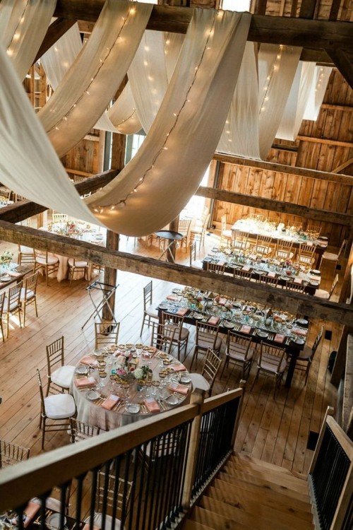 curtains and string lights hanging over the wodoen beams will give a lovely barn wedding venue a veyr cozy and elegant feel