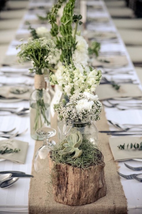 a neutral barn wedding tablescape with a burlap table runner, jars with white blooms and greenery and logs with moss and succulents