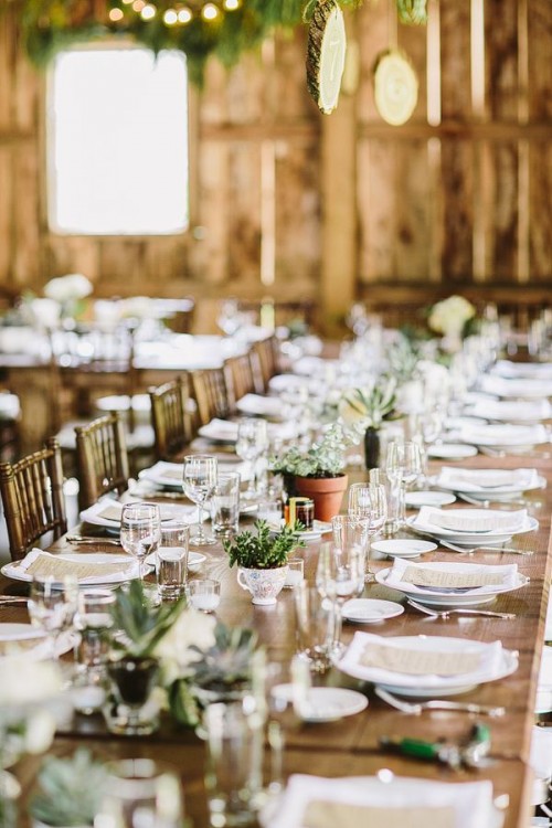 a simple and neutral barn wedding tablescape with a table runner of potted greenery and succulents, neutral linens and menus