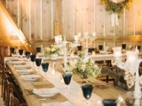 a stylish barn wedding tablescape with an uncovered table, a neutral table runner, greenery wedding centerpieces, candles, blue glasses