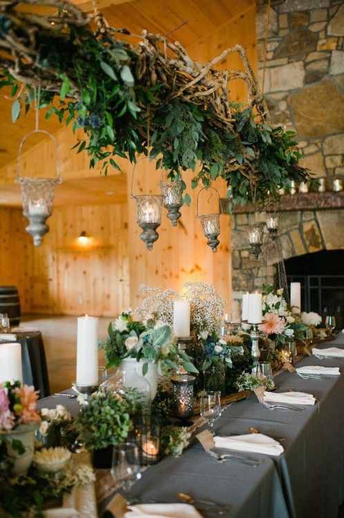 a barn wedding table setting with a grey tablecloth, greenery, white and peachy blooms, candles and an overhead greenery installation with candles