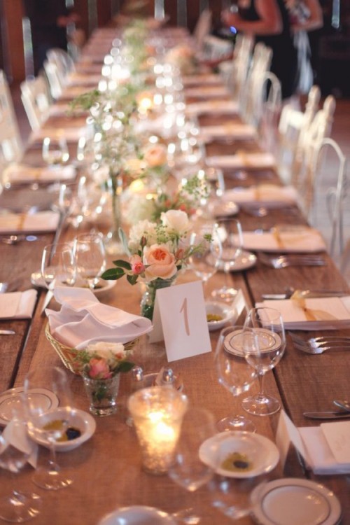 a romantic barn wedding table setting with an uncovered table, a burlap table runner, peachy and white blooms, white linens