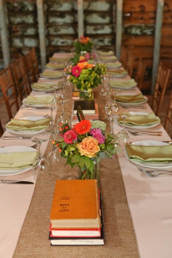 A bright rustic wedding tablescape with a burlap table runner, bright blooms and greenery, vintage books and colorful napkins