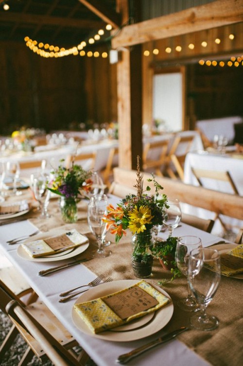 a barn wedding tablescape with a burlap table runner, a colorful floral centerpiece, colorful printed napkins