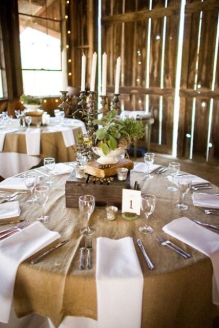 a cool barn wedding tablescape with a burlap tablecloth, white napkins, a box and a book on top and a greenery centerpiece