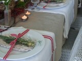 a neutral rustic Christmas tablescape with a burlap tablecloth, striped placemats, a greenery and branch runner, candles, evergreens and red and white ribbons