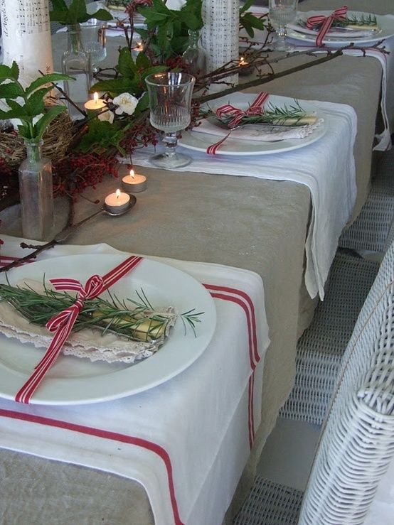 A neutral rustic Christmas tablescape with a burlap tablecloth, striped placemats, a greenery and branch runner, candles, evergreens and red and white ribbons