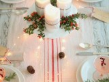a neutral Christmas tablescape with a striped table runner, white porcelain and cranberry cocktails, pinecones, lights and a centerpiece of candles, berries and greenery