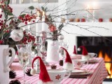 a chic Christmas tablescape in red and white, wiht a plaid tablecloth, branches wit berries and ornaments, pinecones and pompoms, little red caps for each place setting