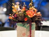 a bright fall wedding centerpiece in a vase wrapped with burlap, bright and dark blooms and artichokes
