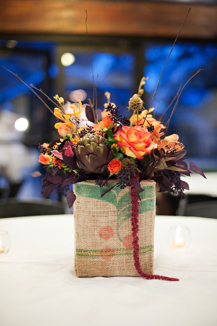 A bright fall wedding centerpiece in a vase wrapped with burlap, bright and dark blooms and artichokes