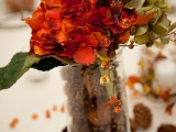 a bold fall wedding centerpiece of a tall glass with pinecones and nuts, bright blooms and foliage and some beads