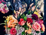 a moody Halloween wedding centerpiece of a black planter, bold blooms, black branches, greenery and candleholders on the branches is very stylish