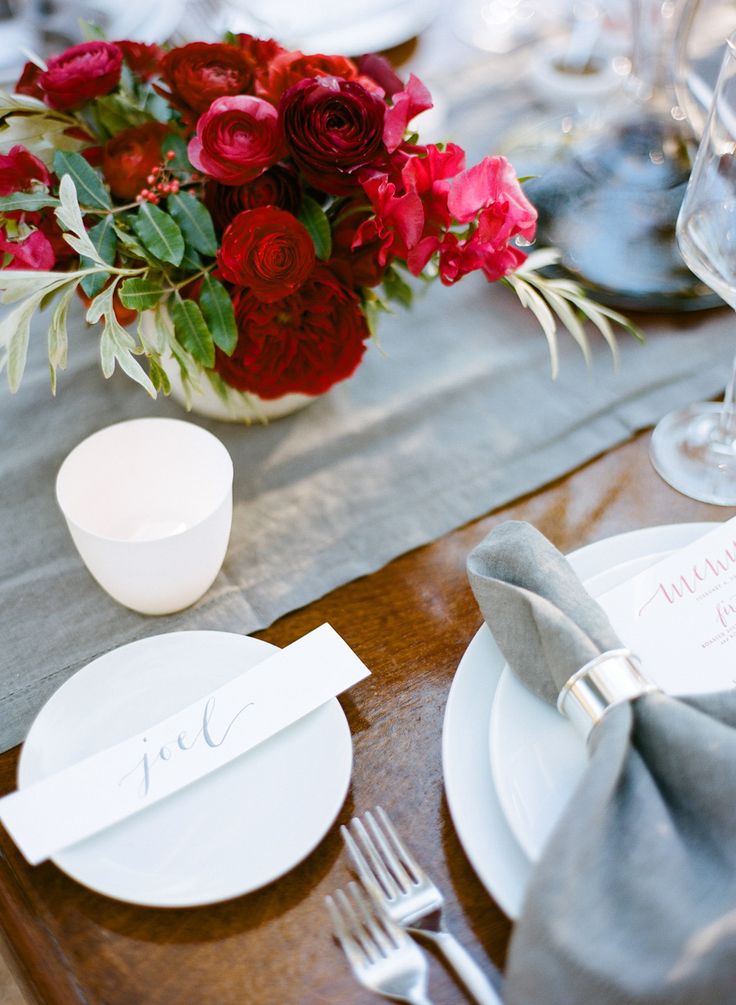 An elegant Valentine's Day wedding tablescape with grey linens, metallic napkin rings, white porcelain, a bold red and burgundy floral arrangement