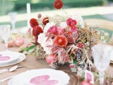 a pretty Valentine’s Day wedding tablespace with an uncovered table, some blush and red blooms, fruits and dried leaves, sheer plates and hearts on each place setting