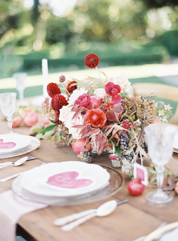 A pretty Valentine's Day wedding tablespace with an uncovered table, some blush and red blooms, fruits and dried leaves, sheer plates and hearts on each place setting