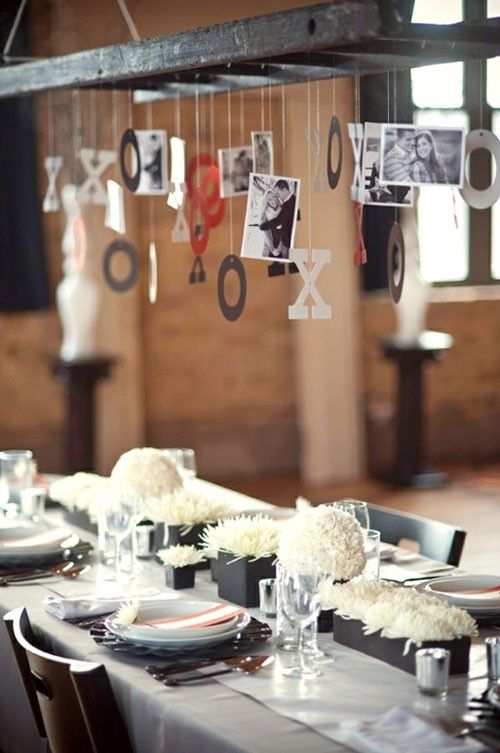 an elegant wedding tablescape with neutral linens, black planters with white blooms, metallic chargers and silver cutlery, an overhead installation with photos and letters for Valentine's Day