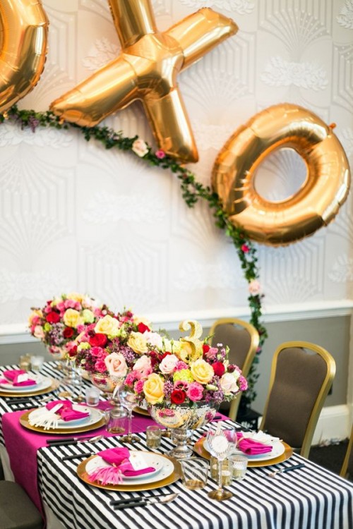 a bold Valentine's Day wedding tablescape with a striped tablecloth, bold fuchsia table runners and napkins, bold floral arrangement, a greenery arch with gold XO letters