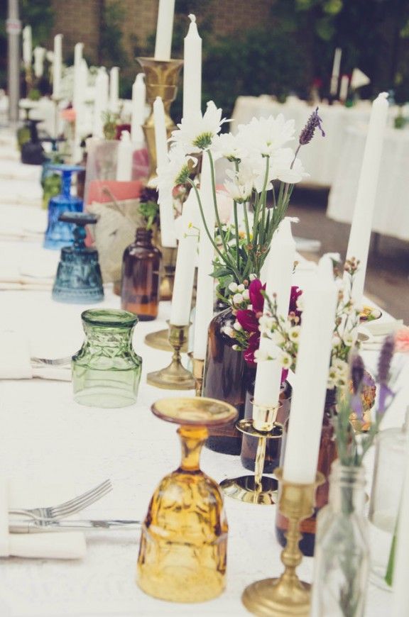 A wedding table setting with candles, dark bottles with blooms, colored glasses for a boho feel