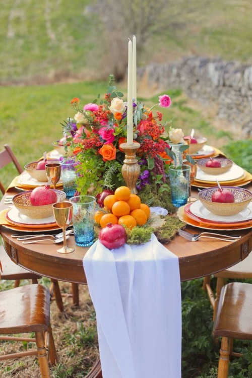 a bright tablescape with a white runner, moss, fruits, a colorful floral centerpiece and gold bowls