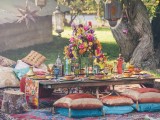a bright picnic setting with a low table, pillows, rugs, a colorful floral centerpiece, colored candles and glasses