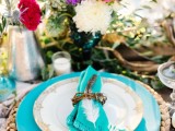 a colorful tablescape with turquoise plates, a wicker charger, bright florals in metallic vases
