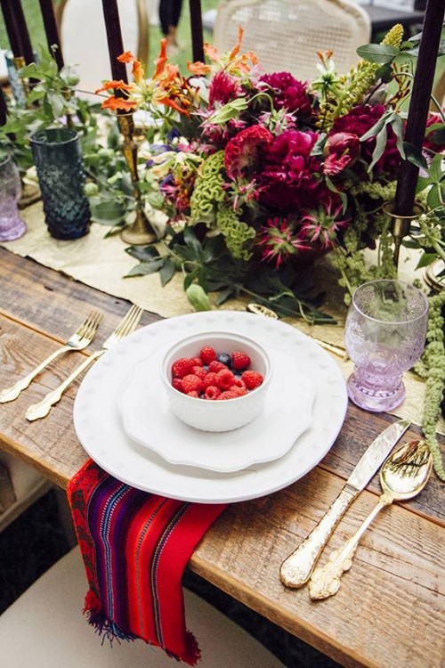 a boho tablescape with a gold table runner, black candles, colored glasses, bright florals and a colorful napkin