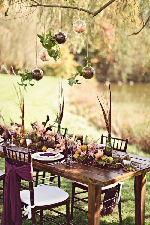 a boho tablescape done with a moss and bloom runner, feathers in bottles, colorful textiles and suspended ornaments with petals