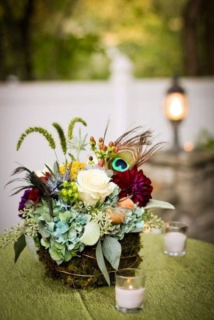 a colorful and quirky wedding centerpiece with greenery, blooms, herbs and peacock feathers