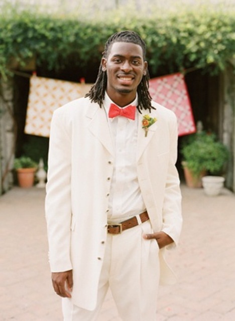 a white suit, a white shirt, a coral bow tie and a bright floral boutonniere for a cool spring or summer groom's look