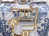 a stylish blue and white wedding tablescape with a printed tablecloth, gold goblets, neutral napkins and a banner on gilded chairs