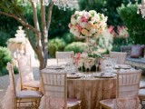 a refined garden bridal shower space with pink chairs and pink ruffle covers, a pink and gold tablecloth, a tall neutral centerpiece and a crystal chandelier