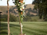 a rustic woodland wedding arch of branches, with greenery and bright blooms and planters wrapped with bark
