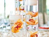 a bright wedding centerpiece of tall glass vases with orange orchids and floating candles is a cool idea for a tropical or beach wedding