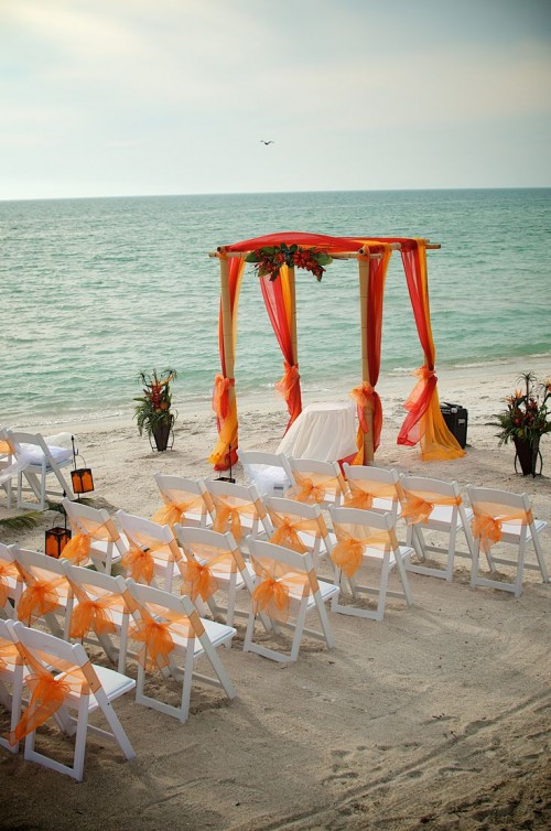 a bright beach wedding ceremony space with a bold orange and fuchsia curtain arch, white chairs with orange ribbons and bold tropical florals is cool