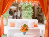 a beach wedding lounge organized with an orange tent and white upholstered furniture, with bold pillows and blooms is a stylish idea for your celebration