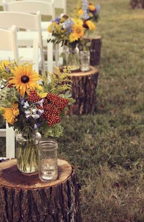 a rustic wedding aisle with tree stumps, floral arrangements and candle jars, simple white wooden chairs