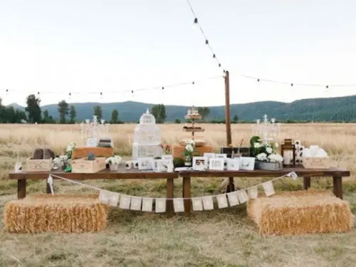 an outdoor wedding reception space with hay as seats, a low and long reception table, string lights over the space and floral arrangements