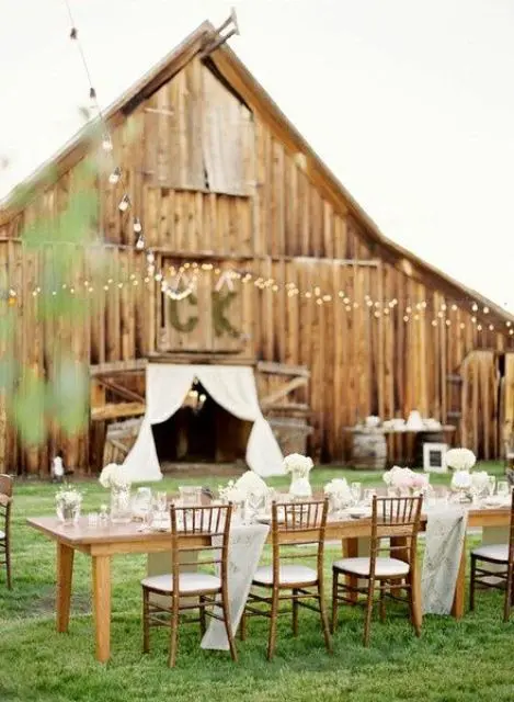 an outdoor barn reception space with long tables, white floral arrangements and string lights over the space