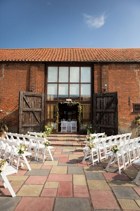 an outdoor barn wedding ceremony space with a greenery arch, simple chairs decorated with greenery and blooms is a cool space