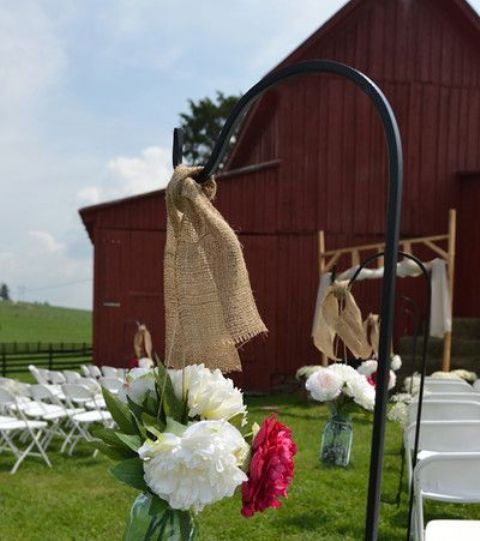 an outdoor barn wedding ceremony space with a wooden arch and burlap, white chairs and floral arrangements and burlap to line up the aisle