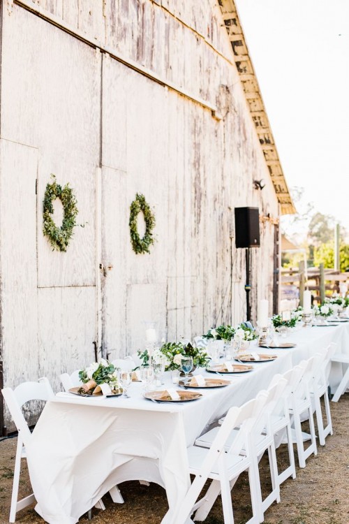 a pretty neutral outdoor barn reception space with a greenery wreath, metal chargers, greenery arrangements and white linens