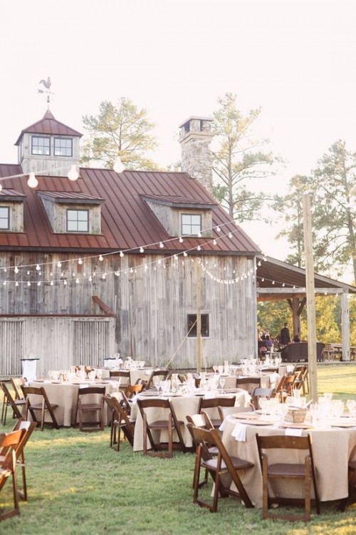 an outdoor barn wedding reception space with round tables, burlap tablecloths, white extiles and string lights over the whole space